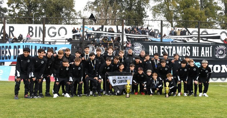 Los chicos de las categorías 2012 y 2014 de Real Pilar, que fueron campeones y subcampeones en la Golden Cup Internacional de Perú, posan con las copas y medallas con la hinchada del Monarca de fondo.