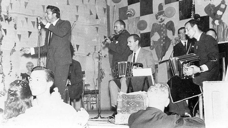 Una orquesta de tangos deleita a los presentes en una imagen de la década del 60. En primer plano se ve a una pareja en pleno baile. .